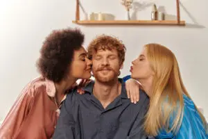 polygamy concept, open relationship, portrait of three adults, multicultural women kissing happy portrait of three adults, women kissing happy man portrayal of threesome