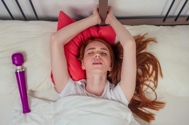 redhaired ginger female person lying on a bed with pink pillows eyes closed from pleasure next to the purple vibrator