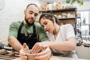 Craftswoman in apron shaping clay bowl with boyfriend and talking while working in ceramic workshop
