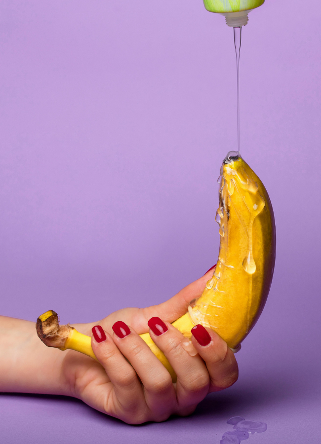 Woman with red fingernail polish holding a banana while lube drips onto the end of it